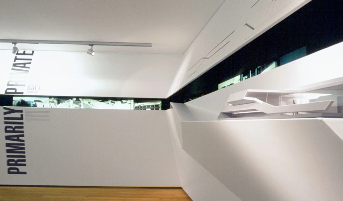 Modern white gallery interior with an angular geometric sculpture on a pedestal; vertical PRIMARILY text on the left wall