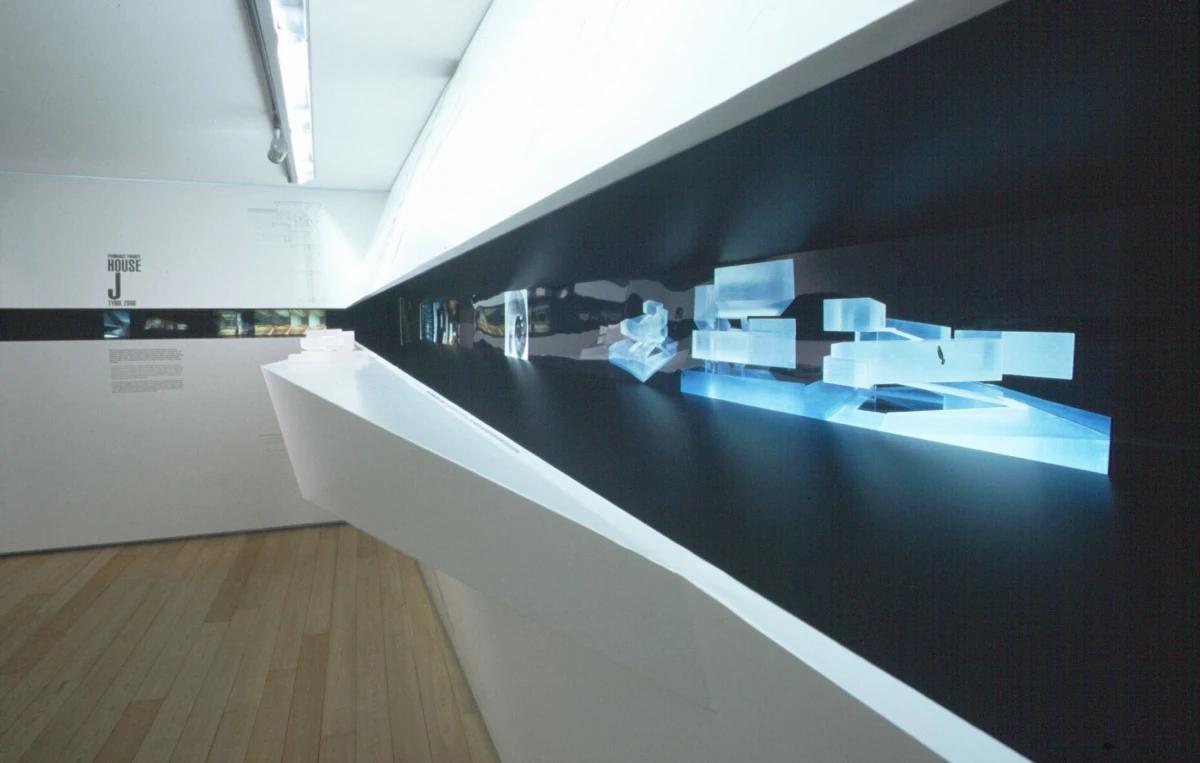 Contemporary art gallery interior featuring a white angled display platform against a black wall with glowing blue translucent blocks.