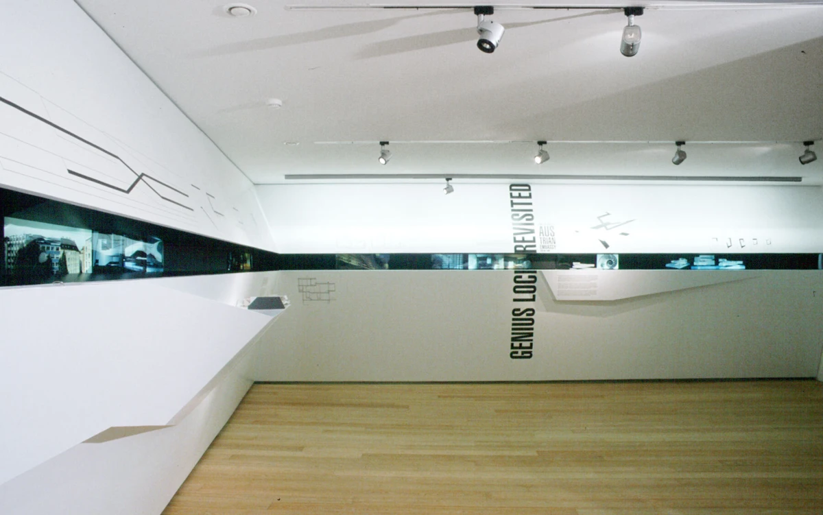 Minimalist art gallery with white angular display, black mirrored wall strip, wooden floor, and vertical 'GENIUS LOCI' text on center wall.