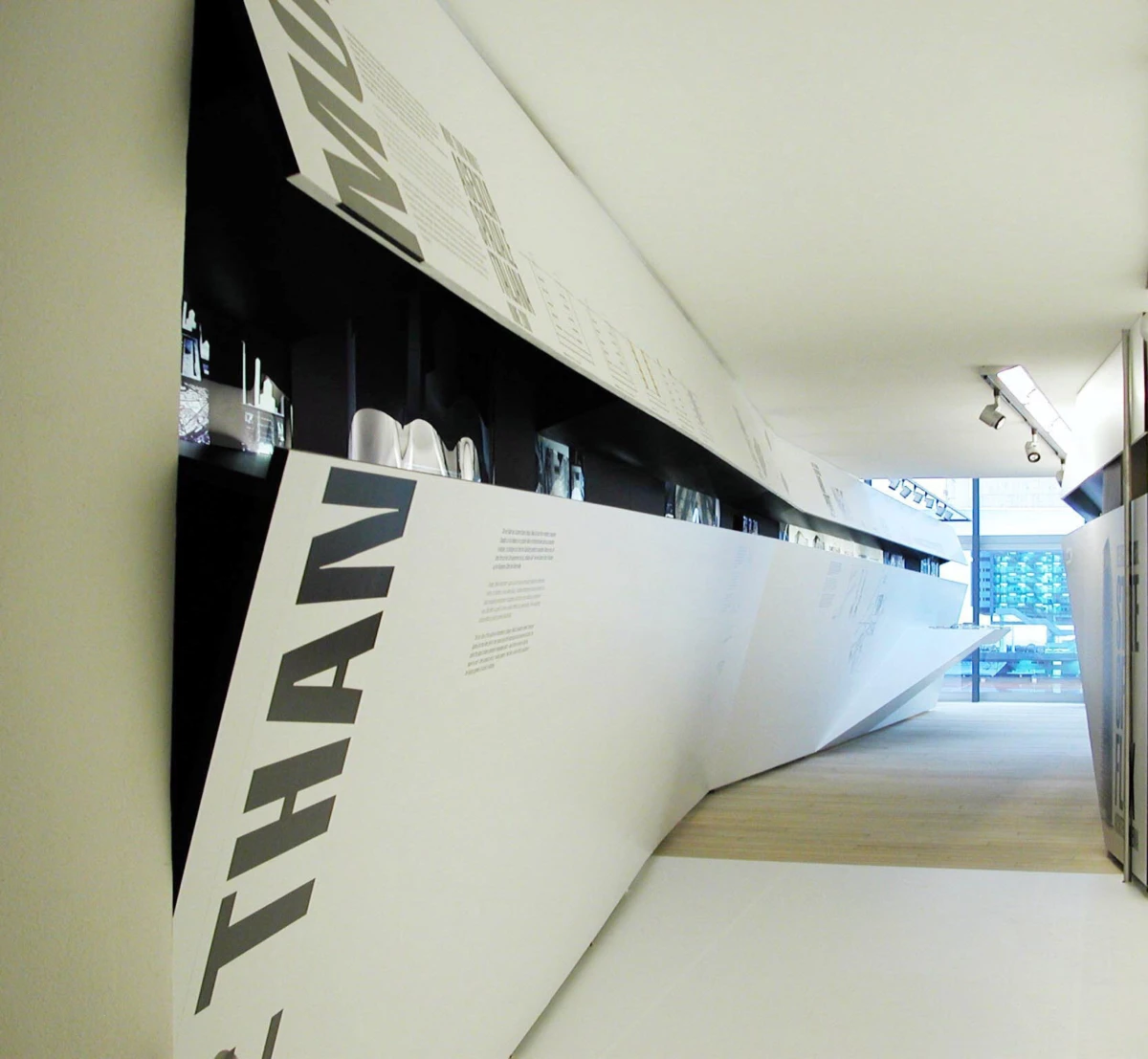 Modern museum hallway with a slanted white display wall, black interior shelves, and faint text panels.
