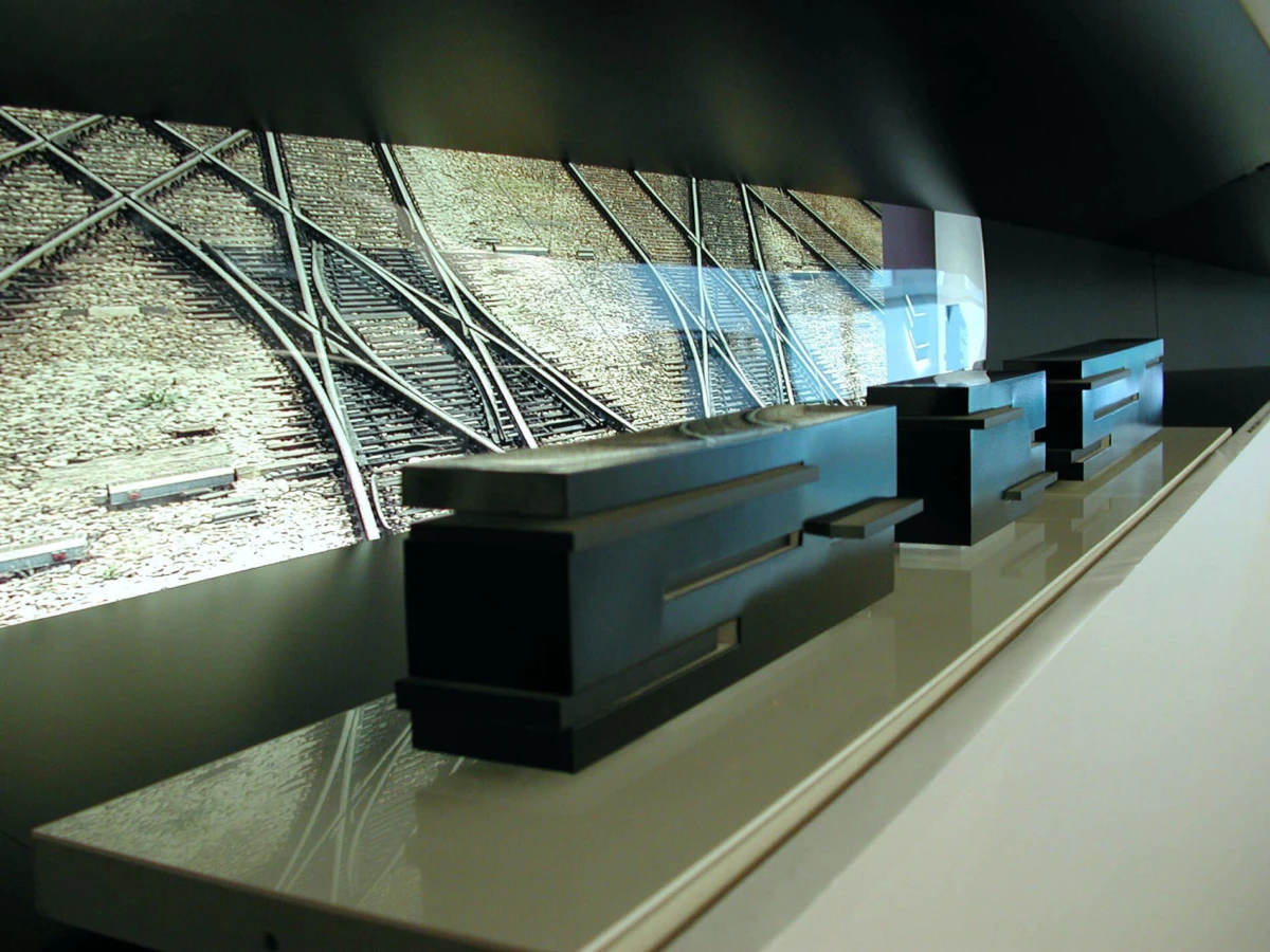 Abstract black rectangular prisms arranged on a glossy white shelf, with a railway-tracks photograph backdrop.