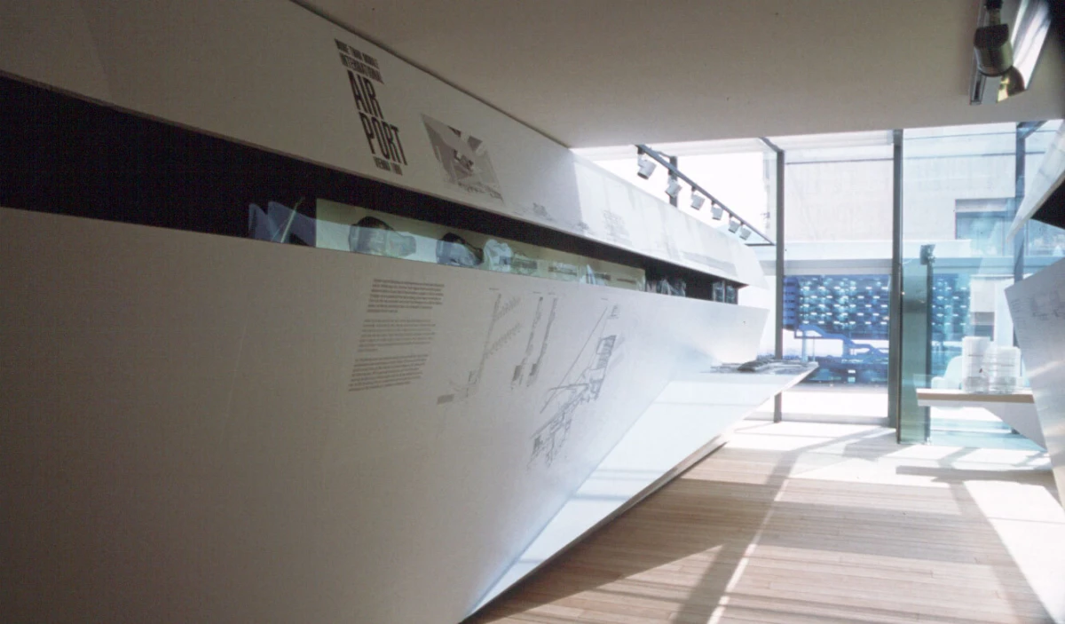 Modern gallery interior with a long white exhibit wall, glass panels, wooden floor, and daylight streaming through glass doors.