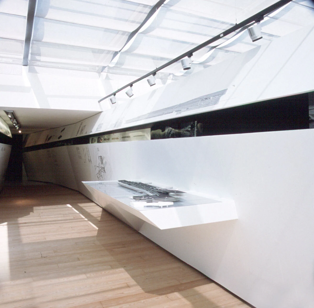White minimalist gallery interior featuring a diagonal wall-mounted display shelf with metal objects under track lighting.