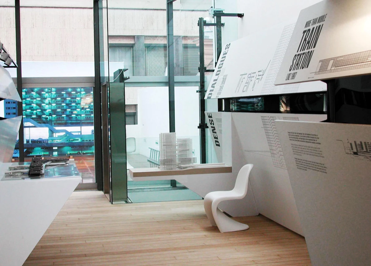 White angular display panels in a modern exhibition; curved chair and brochure table, with glass walls revealing blue seating.