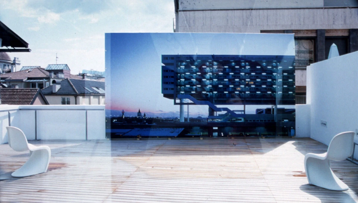Blue glass-panel sculpture on a rooftop terrace, flanked by white modern chairs, with city skyline in background.