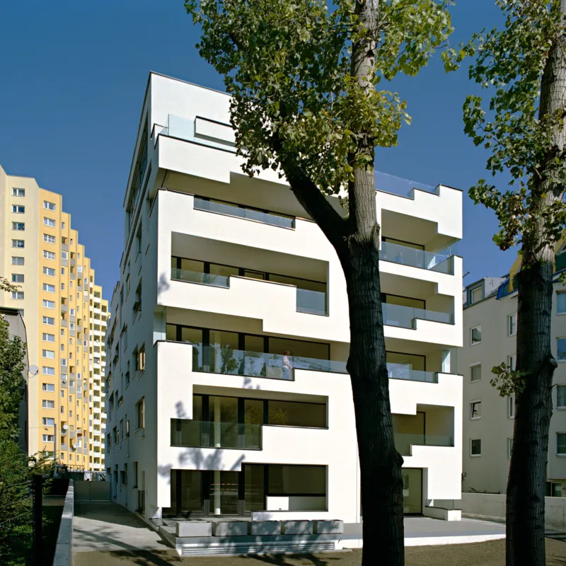 White modern apartment building with staggered balconies framed by large trees on a sunny urban street.