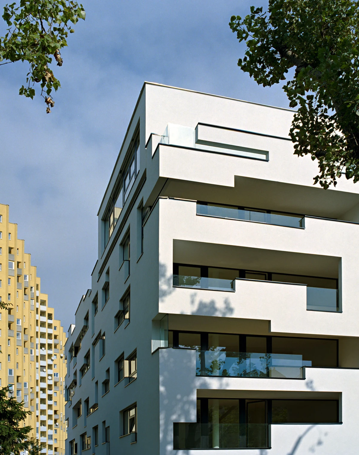 Modern white apartment building with staggered balconies and glass railings in an urban setting under a blue sky.