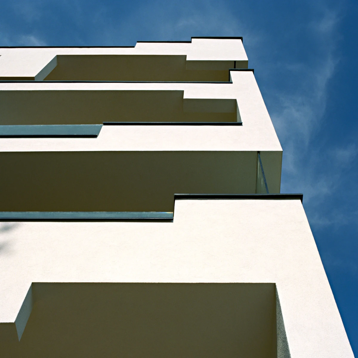 Low-angle view of a modern white apartment building with stacked cantilevered balconies against a blue sky.