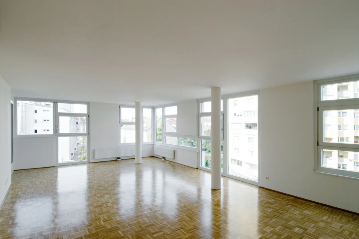 Bright empty living room with parquet floor, white walls, and large windows; a white column near the center.