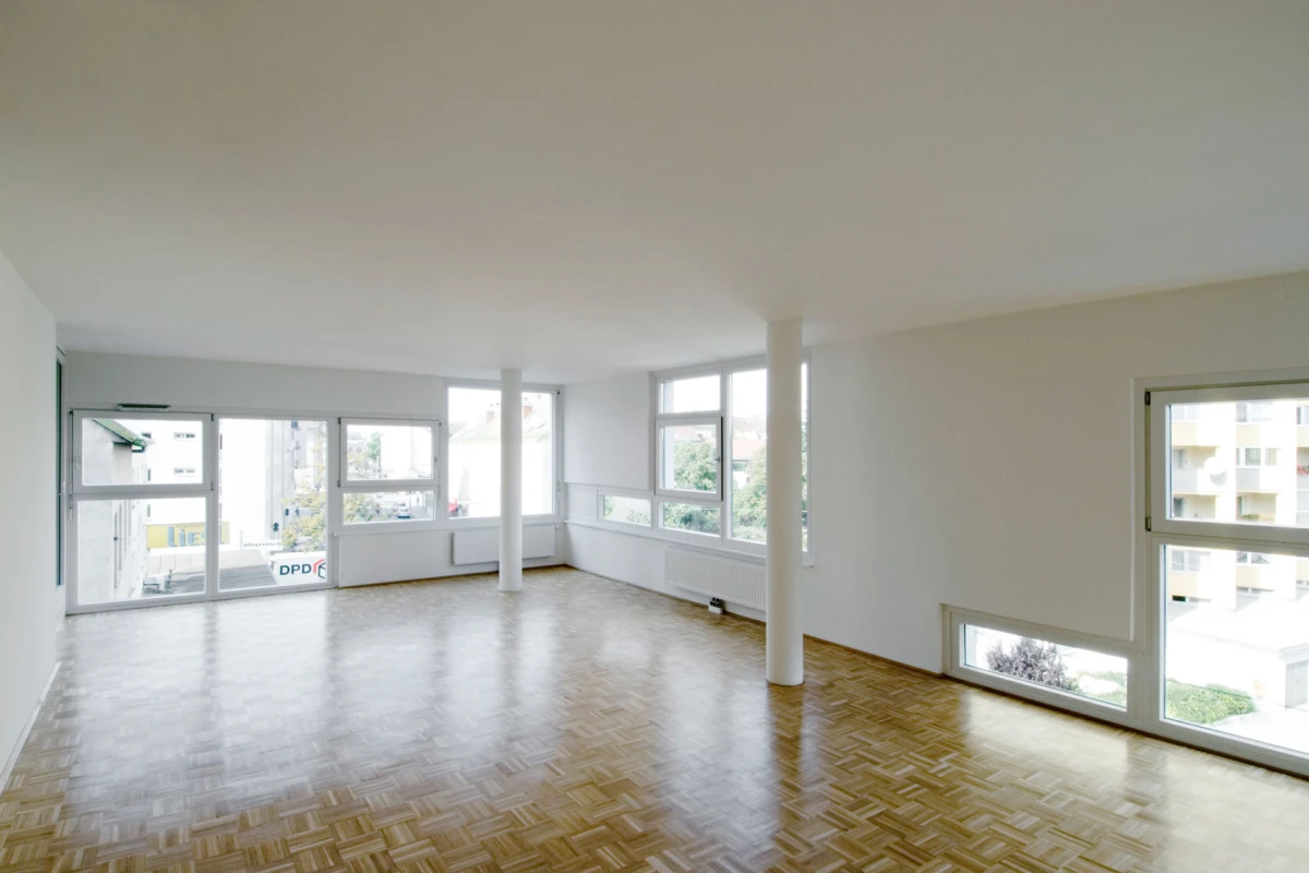 Bright unfurnished living space with parquet floor and white walls, featuring large windows along two walls and white columns.