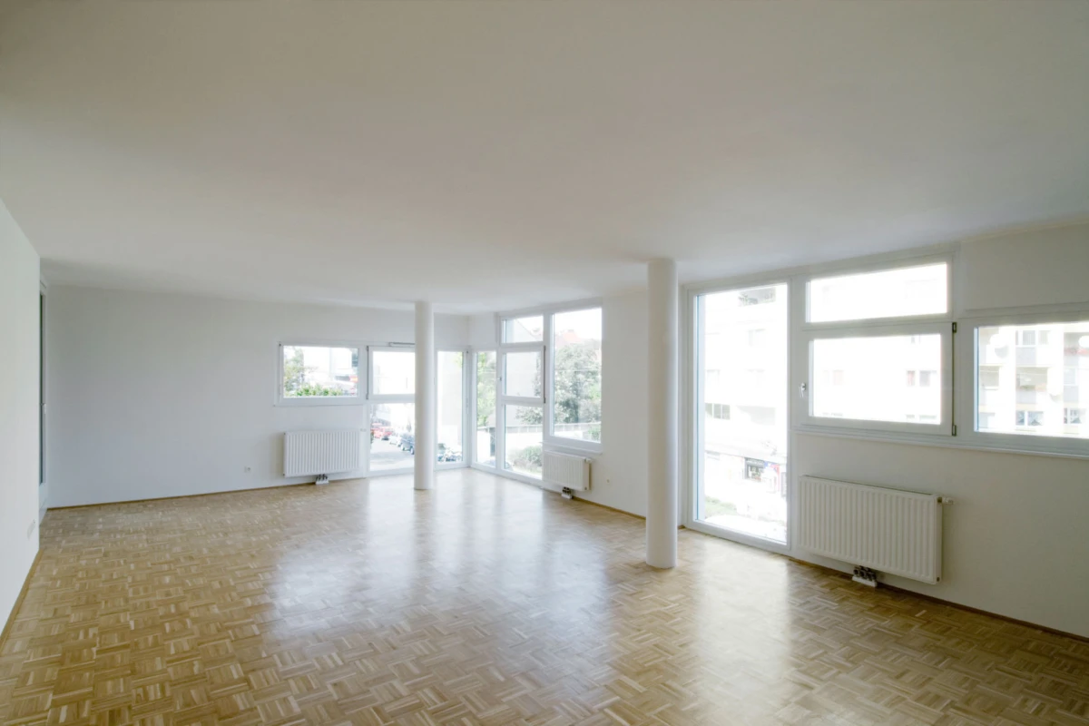 Empty living room with white walls, light parquet floor, large windows and glass door, and white radiators.