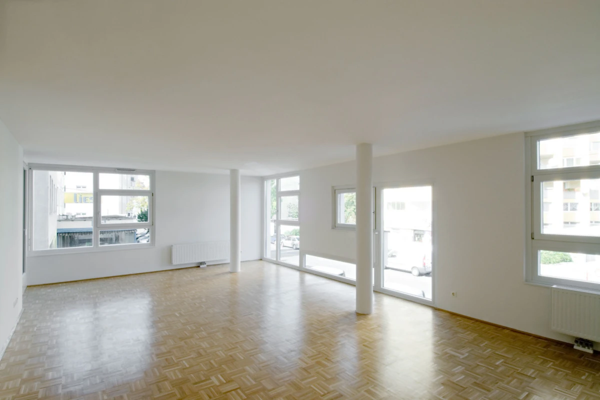 Empty living room with parquet floor, white walls, a central column, and large windows streaming daylight.