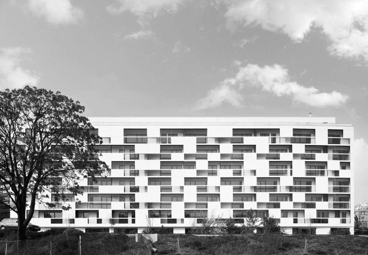White modern apartment building with irregular, stacked balconies; urban lot, grassy foreground, large tree on the left, cloudy sky.