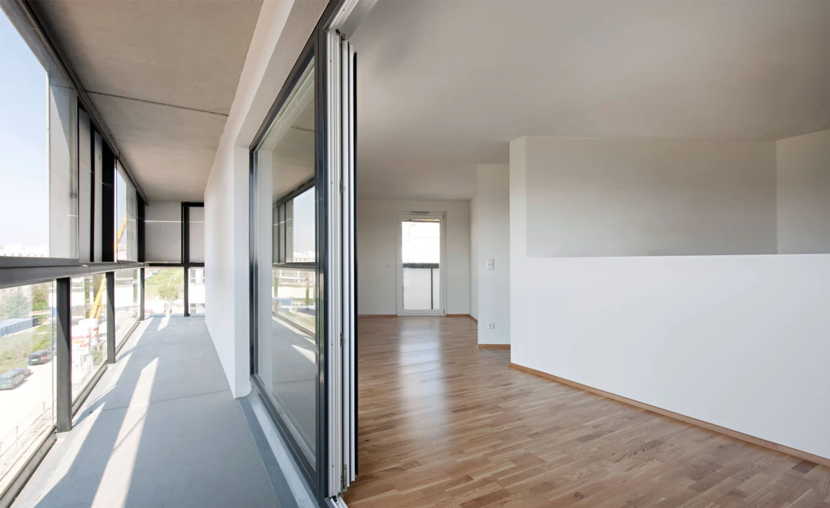 Empty modern apartment living area with light wood flooring, white walls, and sliding glass doors opening to a sunlit balcony.