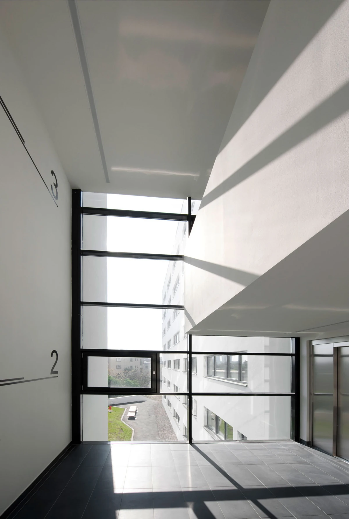 Modern building corridor with large glass wall and black window frames; sunlight creates striped shadows on dark tile floor; view of courtyard outside.