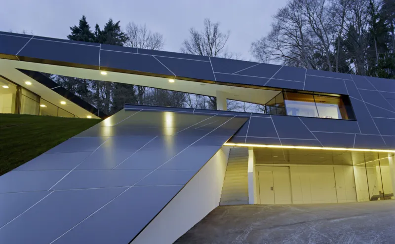Modern angular residence clad in steel-grey panels with an illuminated entry and garage, set on a hillside at dusk.