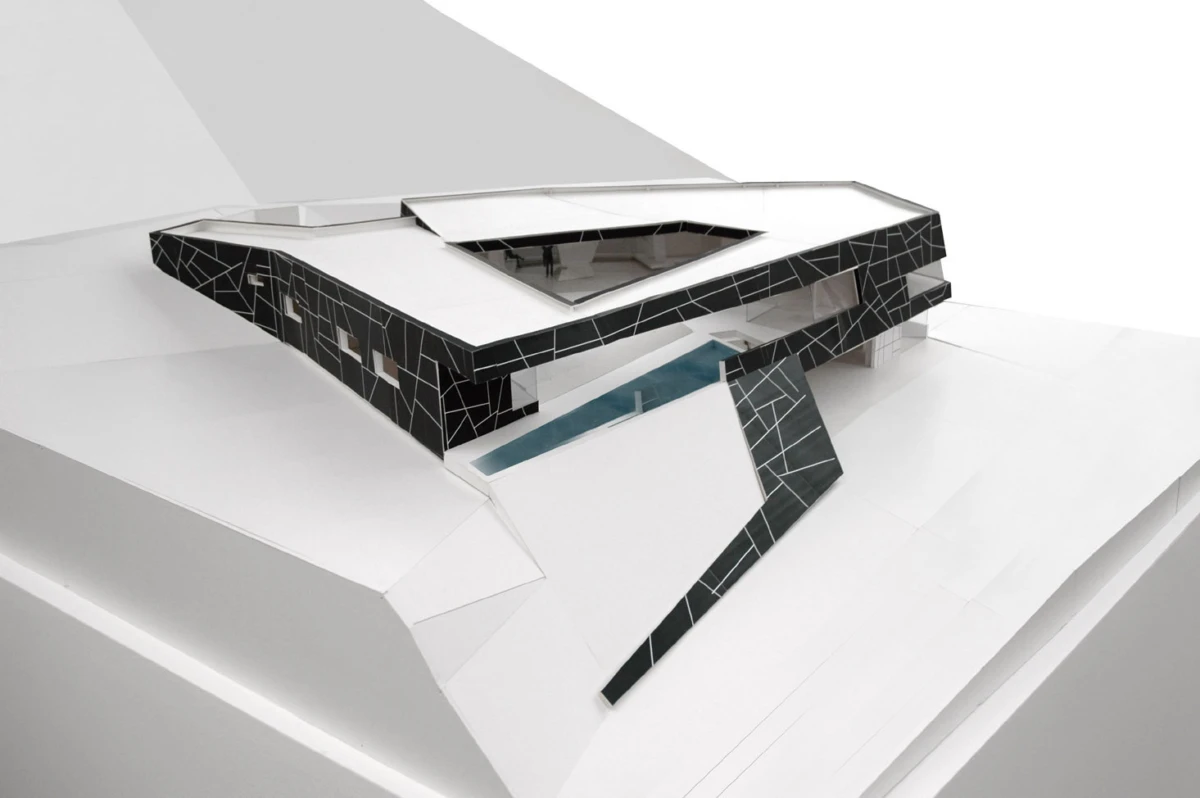 Architectural model of a modern black-mosaic house with an angular roof, perched above a blue rectangular pool on a white display platform.