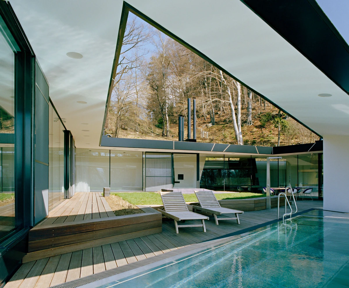 Modern glass-walled residence with a rectangular pool; two wooden lounge chairs on a wooden deck, hillside and trees beyond.