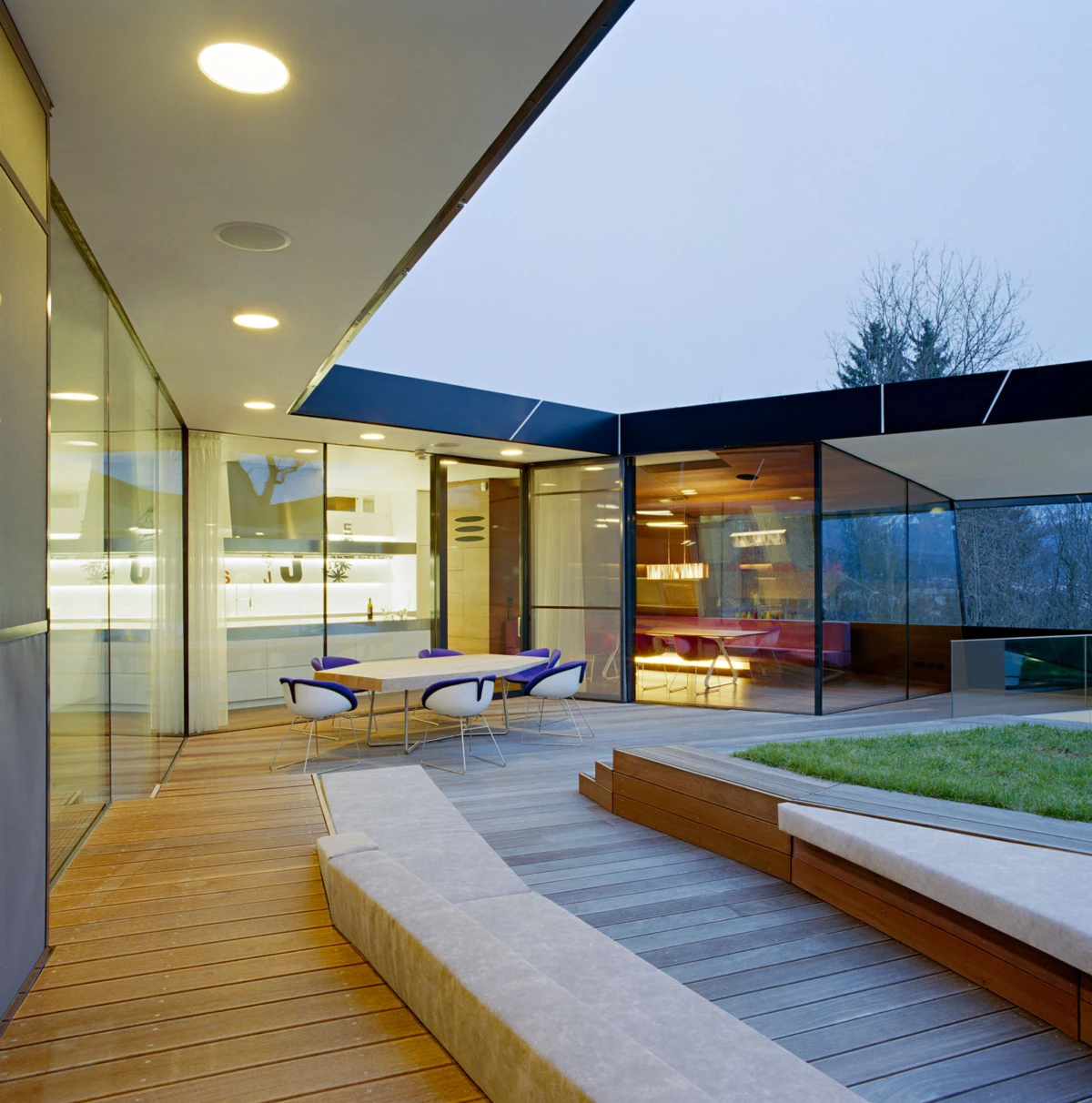 Empty outdoor dining area on a modern glass-walled patio with a wooden deck and blue chairs.