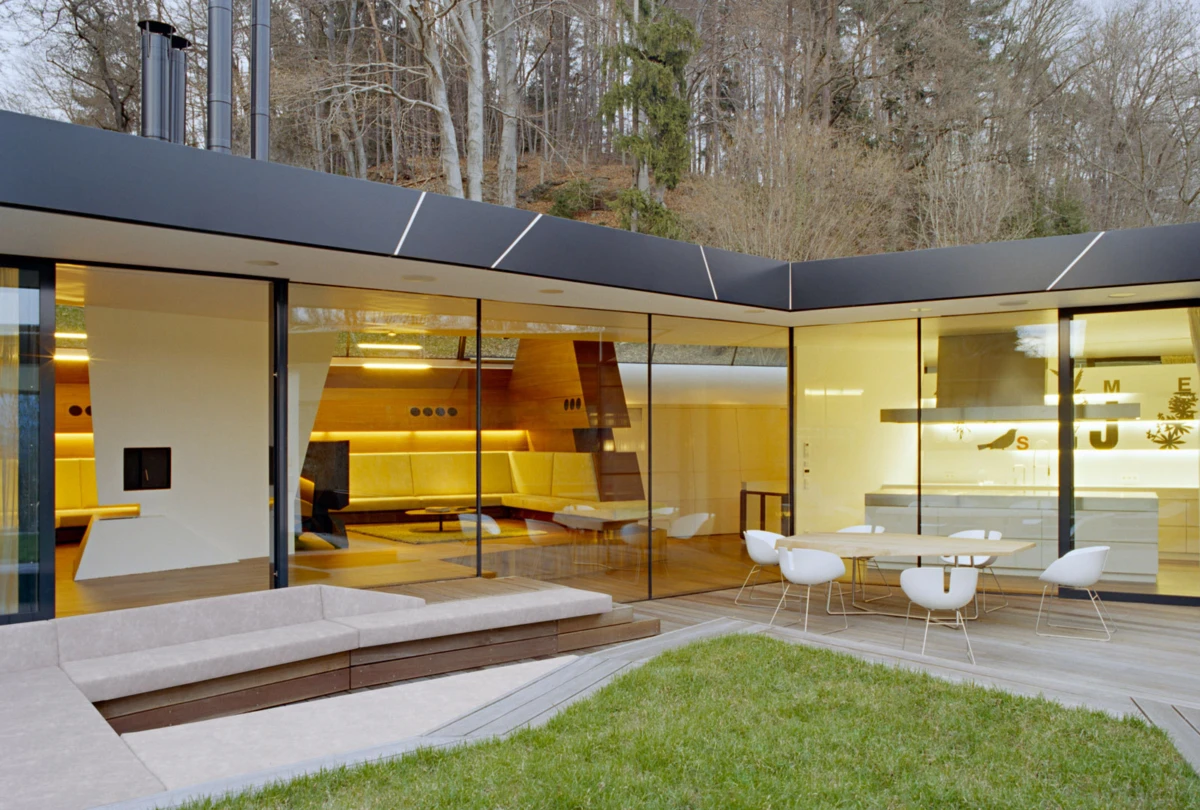 Modern glass-walled home with warm interior; outdoor patio with white table and chairs on a wooden deck, lawn in foreground.