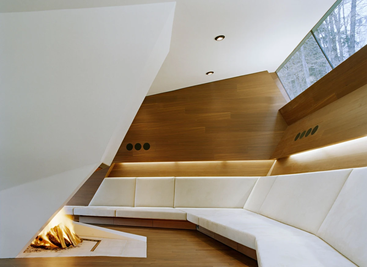 Modern living room with white sectional sofa along a wood-paneled wall, a gas fireplace, and angled white walls; large window reveals trees outside.