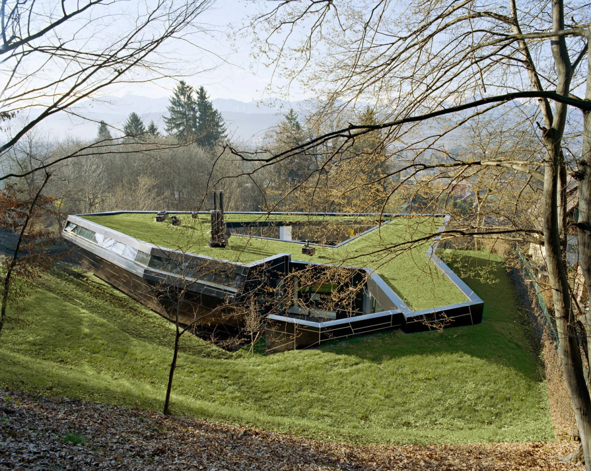 Green-roofed modern glass-and-steel building on a grassy hill, framed by leafless trees.