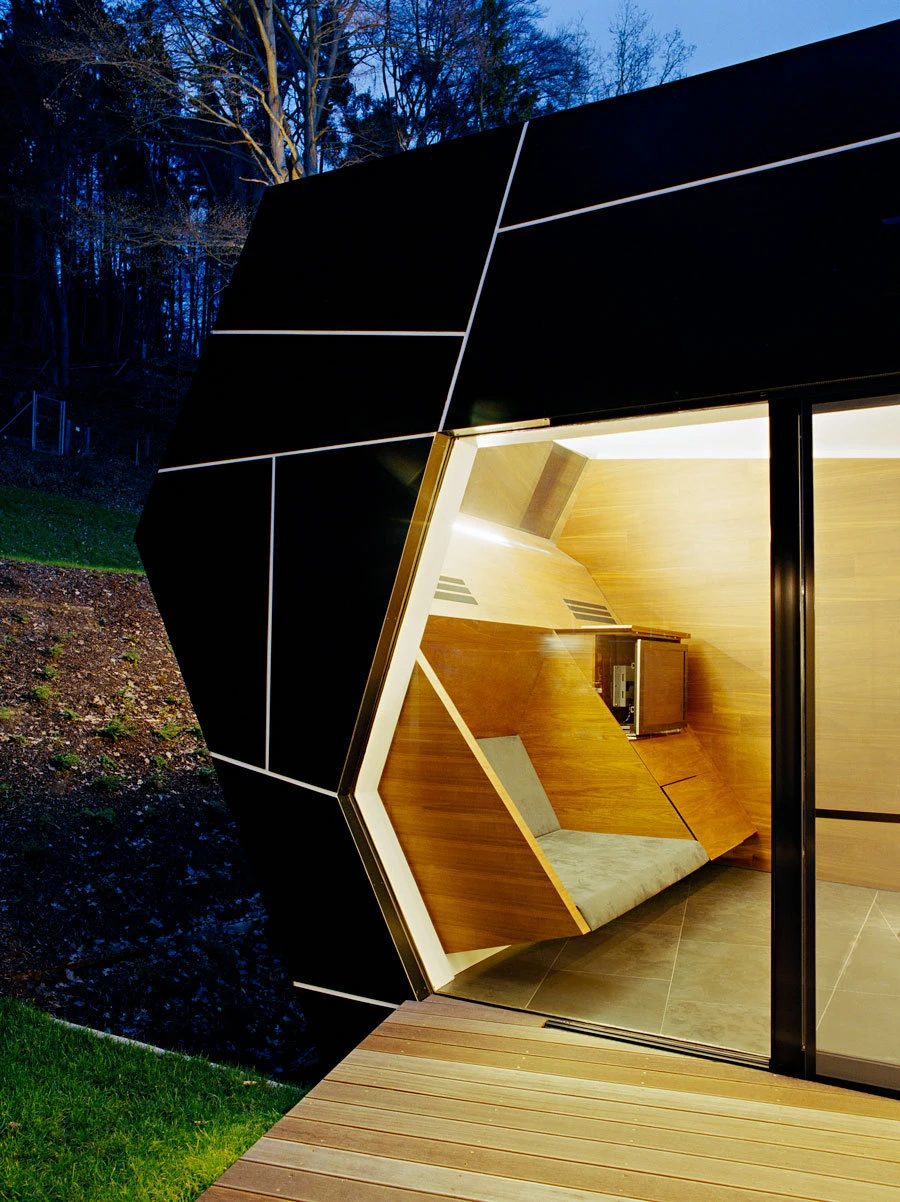 Polygonal black pod with glass entry; warm wooden seating nook inside; exterior on a wooden deck beside a forest at dusk.