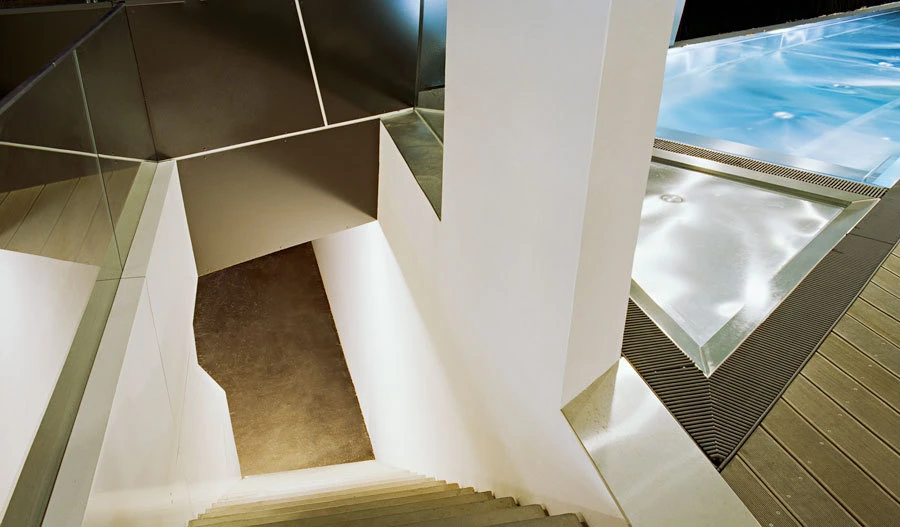 Modern white stairwell with glass railing descends beside a blue indoor pool.