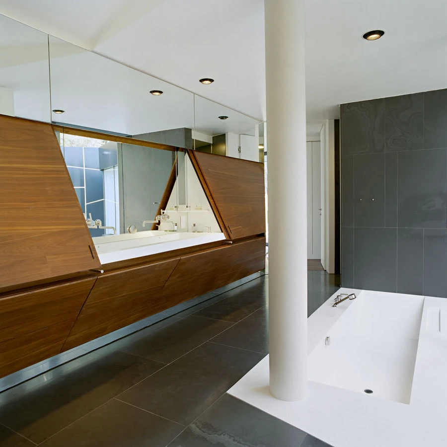 Modern bathroom featuring a walnut angled vanity with large mirrors, white countertop sinks, and a white freestanding tub next to a cylindrical column.