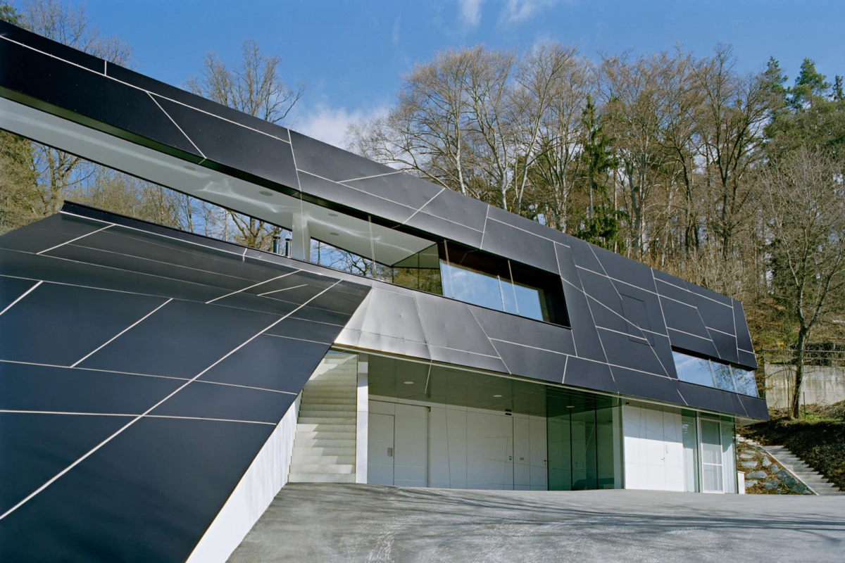 Modern angular building clad in dark metal panels with glass entrances; concrete driveway and forested hillside behind.