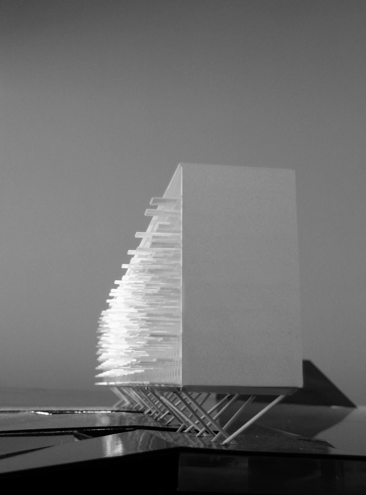 Abstract architectural sculpture: tall rectangular block with stacked translucent lamellae on angled supports, placed on a dark pedestal.