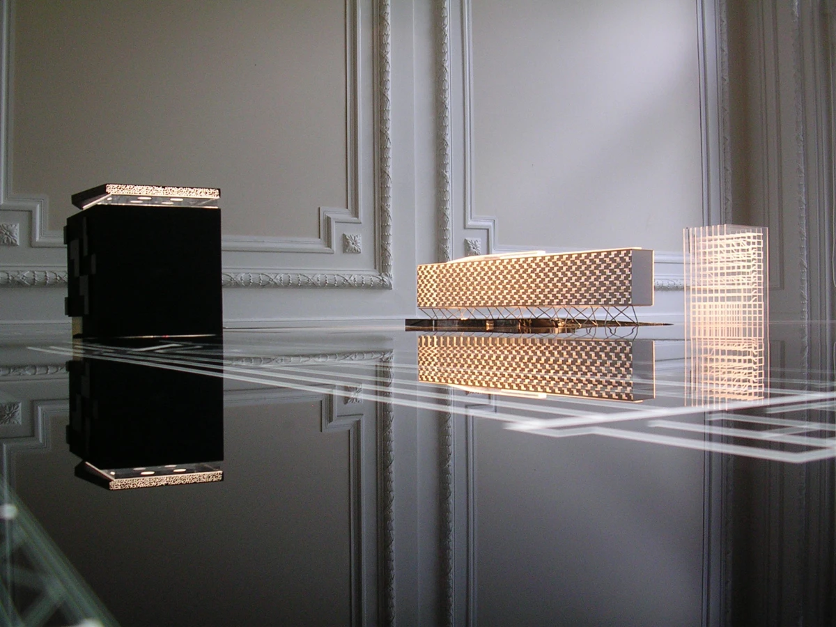 Three architectural scale models on a reflective surface in an ornate room: a black cuboid, a gold lattice bridge, and a tall grid tower.
