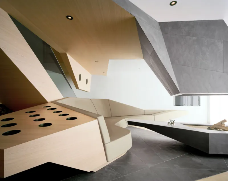 Modern angular interior with beige upholstered seating, wooden and gray concrete planes, and a perforated wood divider.