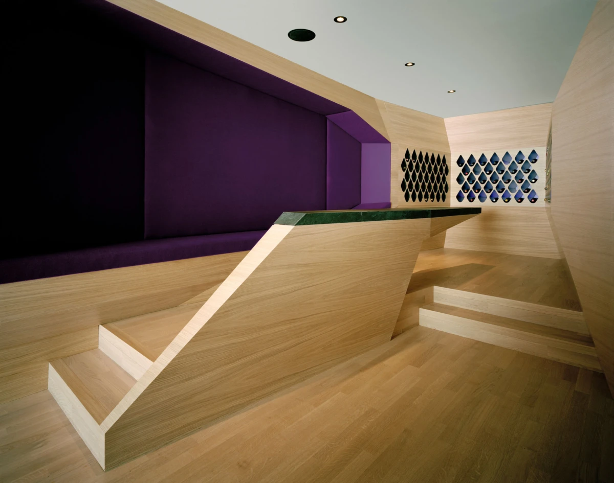 Interior scene with light wood staircase ascending to a purple seating alcove; beige walls with wine racks.