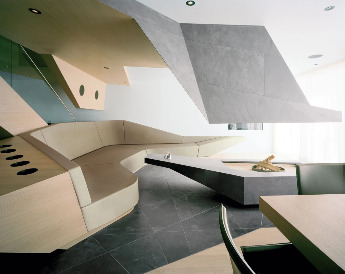 Contemporary interior with beige curved sectional sofa, angular grey ceiling beams, and a low concrete coffee table in a bright lounge.