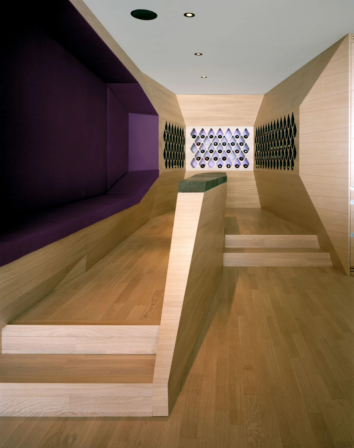 Wooden stepped ramp to a raised platform in a modern interior; purple seating along the left wall and a diamond-patterned wall at the far end.