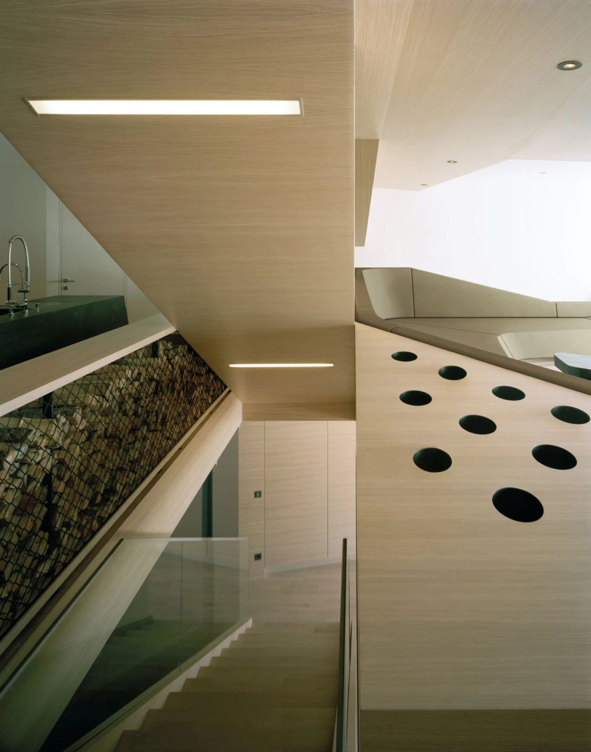 Modern interior with wooden paneling, a glass-railed staircase, and a curved wall with black circular perforations under ceiling lights.