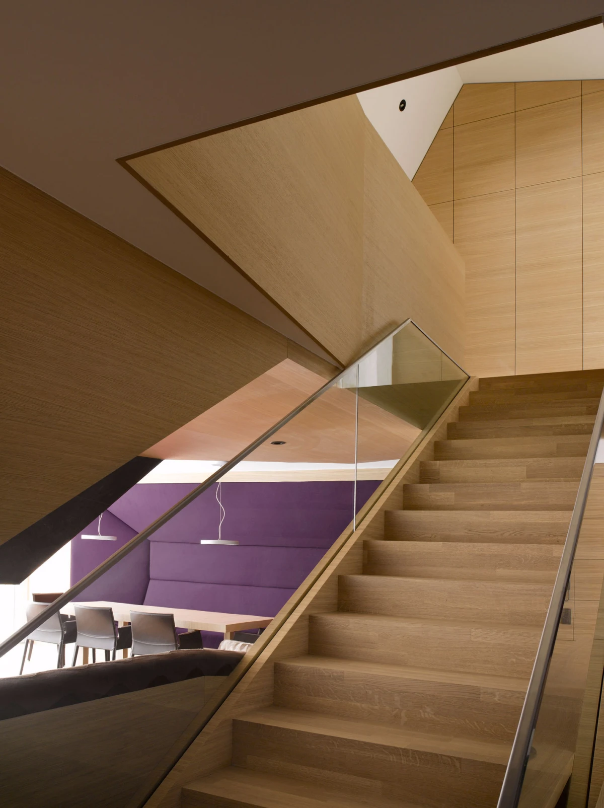 Wooden staircase with glass railing ascending in a modern interior; purple lounge wall and dining chairs visible beyond the glass.