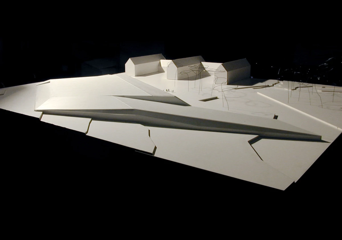 White architectural scale model of a modern complex with sharp-angled roofs on a sloped base; sparse wire trees against a dark background