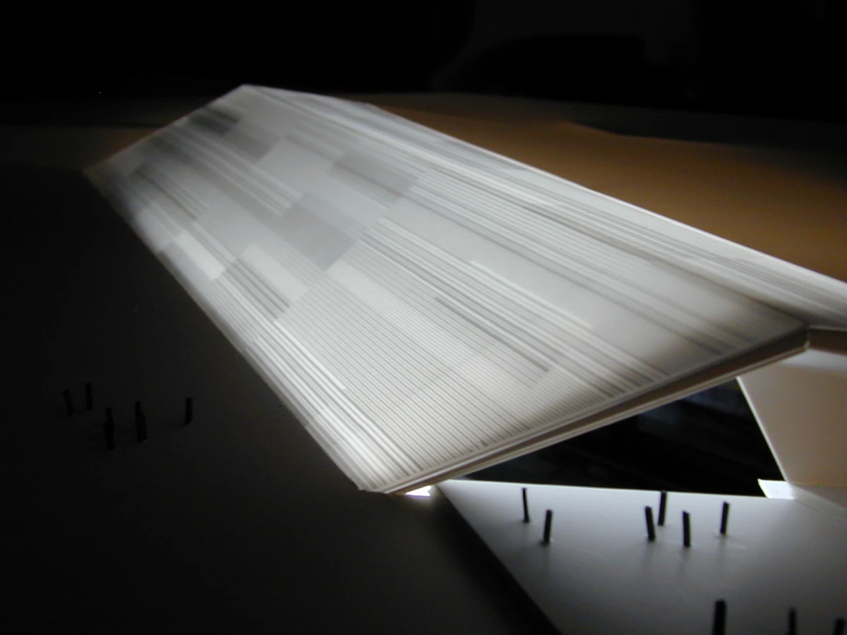 Tilted white architectural paper model with layered panels over a dark base with pins.