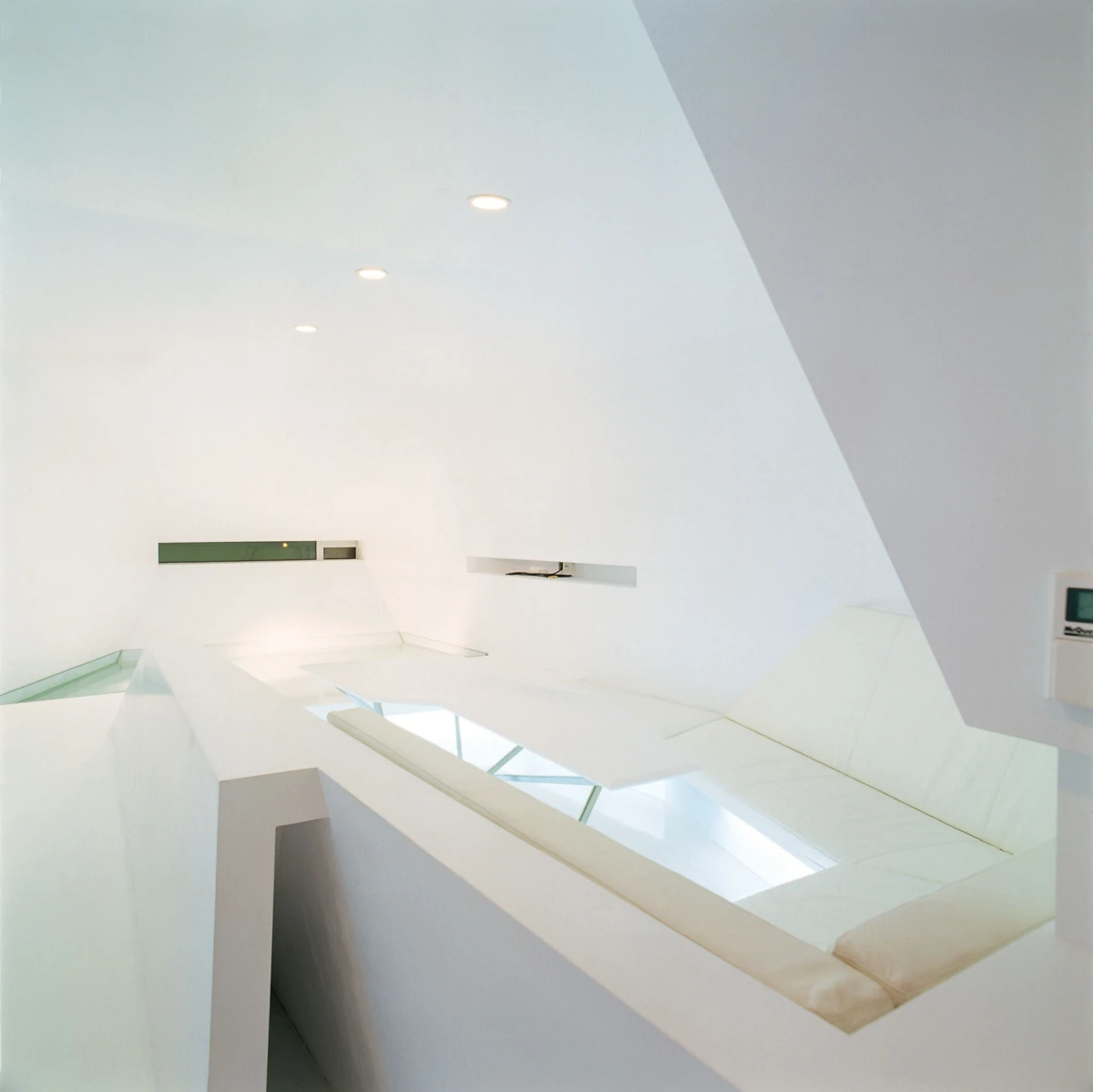 Minimalist white architectural interior with angular walls, glass skylights, and a cushioned white ledge.