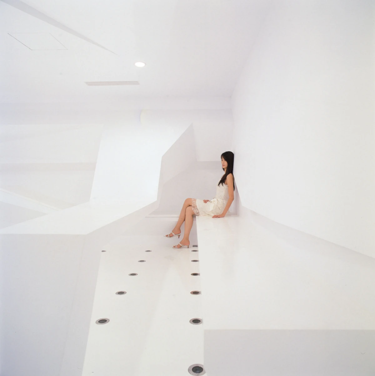 Woman in a white dress with white high-heeled sandals sits on a white platform in a minimalistic, all-white interior.