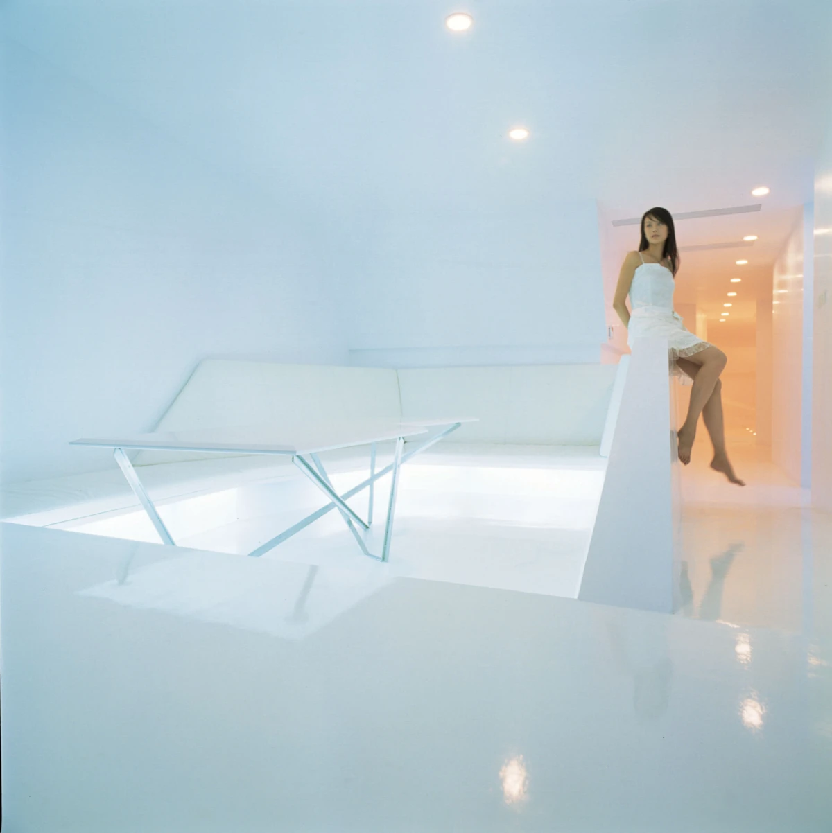 Person in white dress stands on a white angular ledge in a bright minimalist white room with a hallway in the background.