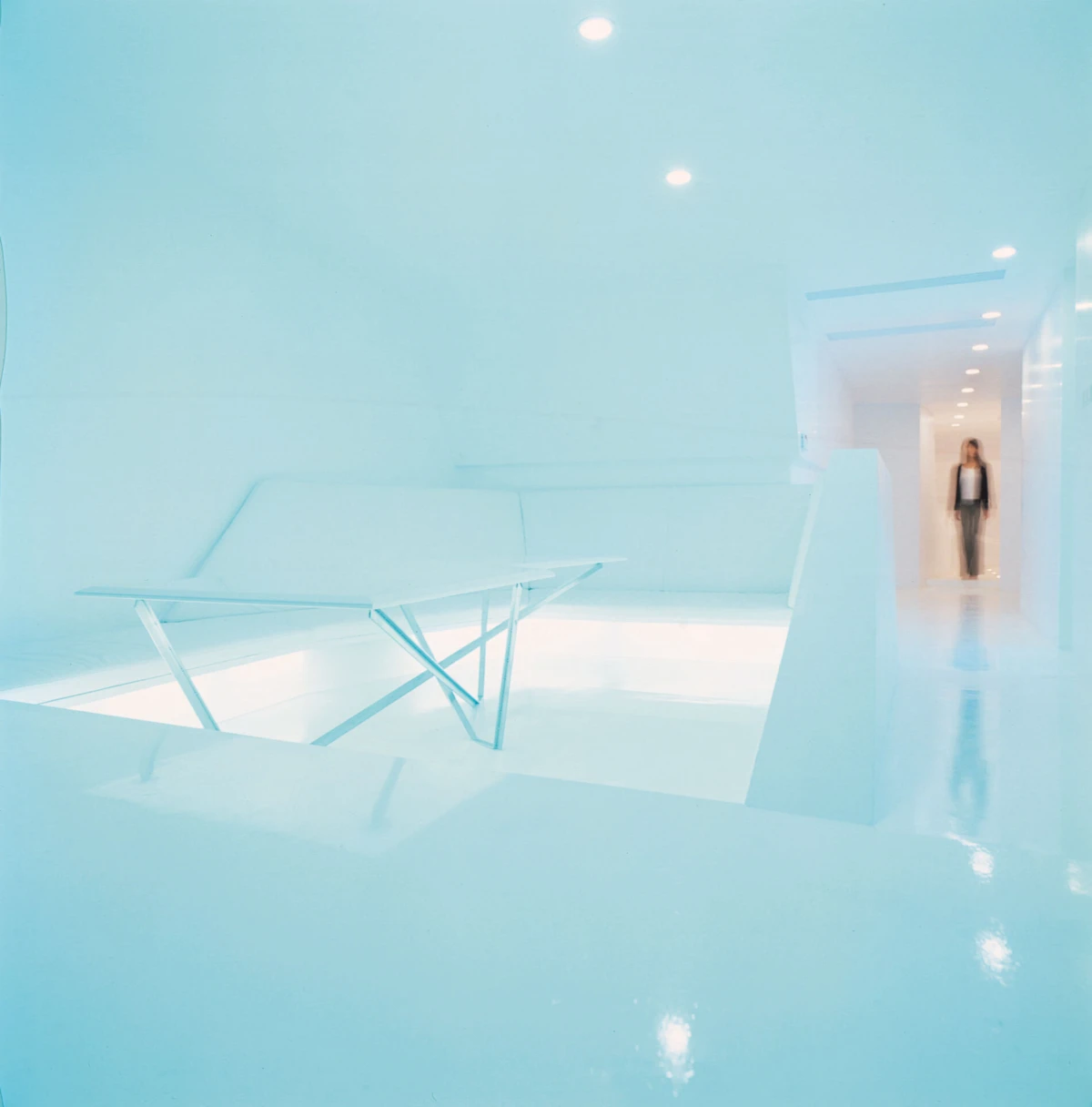 Minimalist white interior with a folding table; a person stands at the end of a bright corridor.