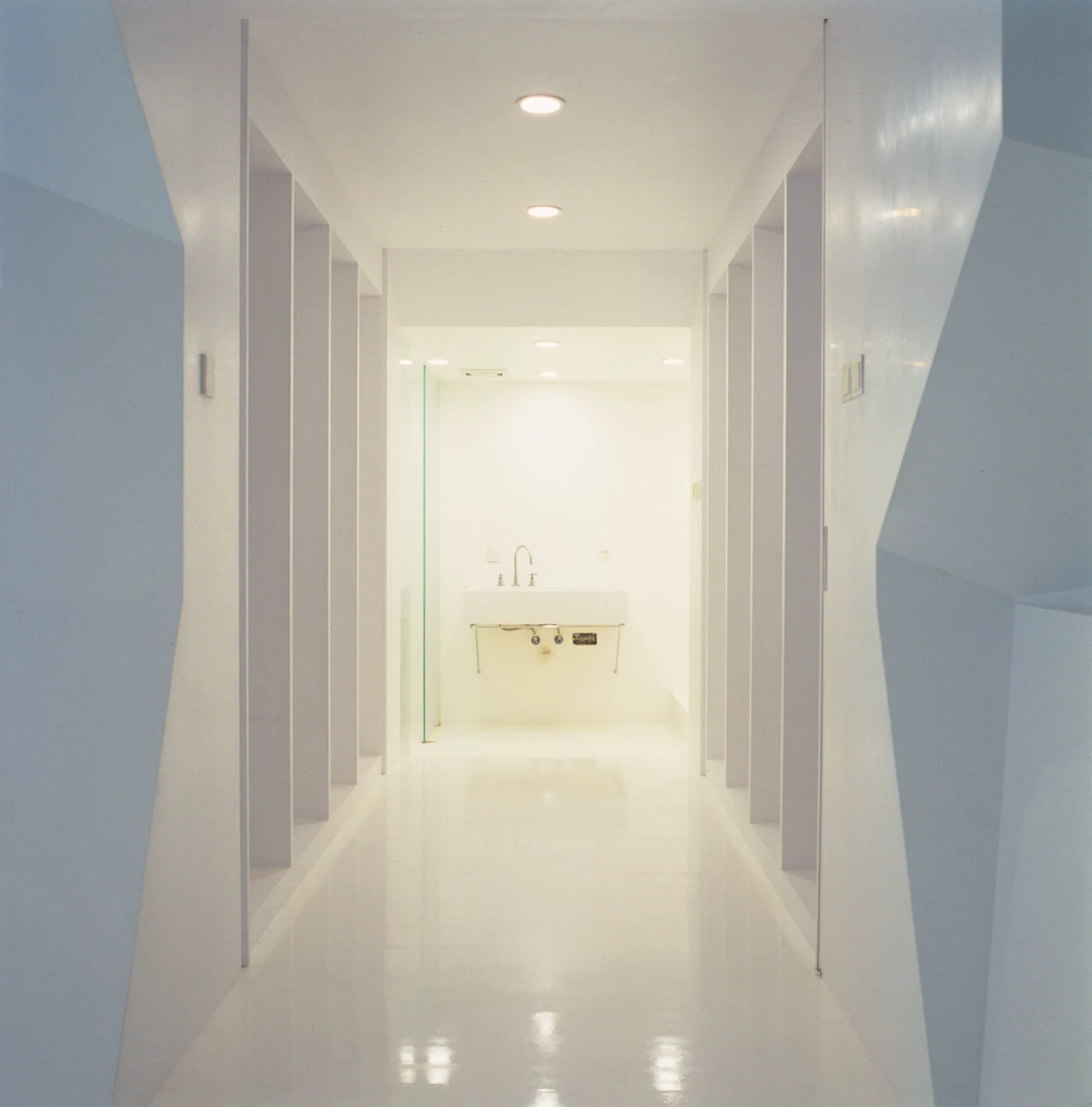 Bright white corridor with recessed ceiling lights and side alcoves, ending with a white sink on the far wall.