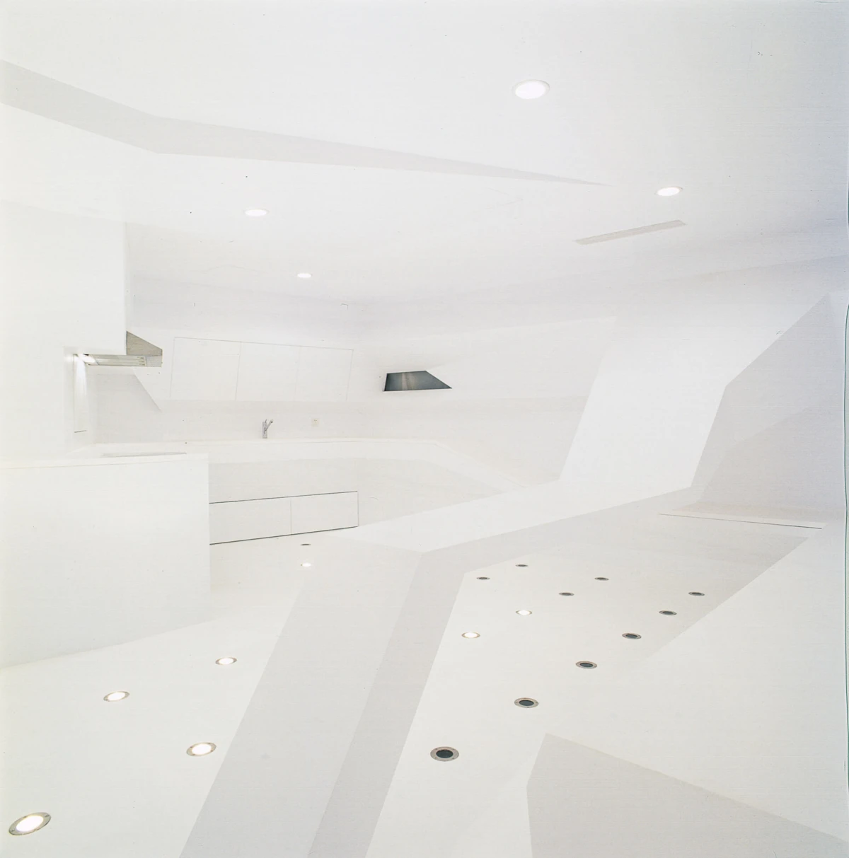 Empty white minimalist kitchen with angular countertops, built-in cabinets, and recessed ceiling lights.