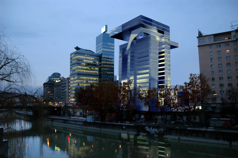 Ultramodern blue-glass skyscraper with an overhanging top beside a river, reflecting in the water, with surrounding buildings at dusk.