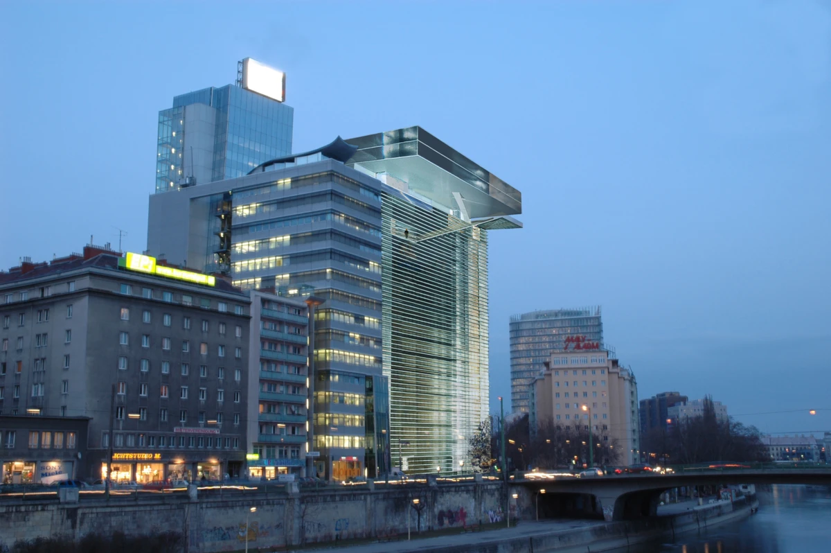 Evening cityscape with a glass high-rise featuring an overhanging cantilever, beside older riverfront buildings.