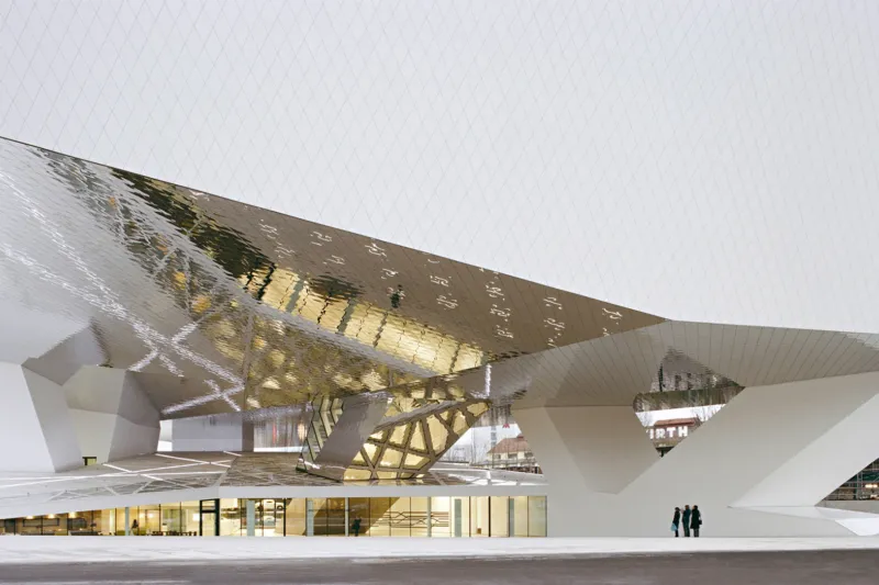 Futuristic white, angular building with reflective metallic panels; two people stand at its base.