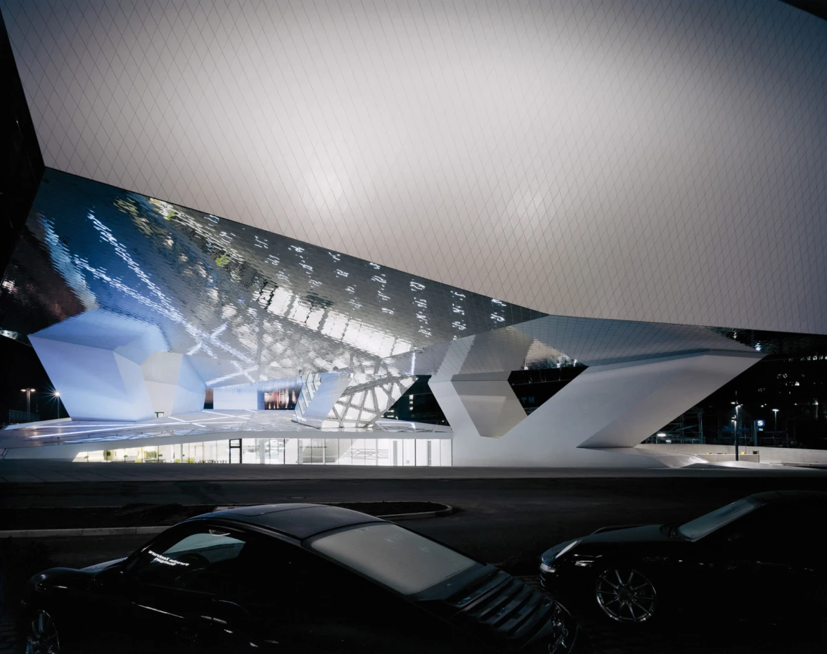 Night view of a futuristic white geometric building with a mirrored roof, glass walls, and parked cars in the foreground.